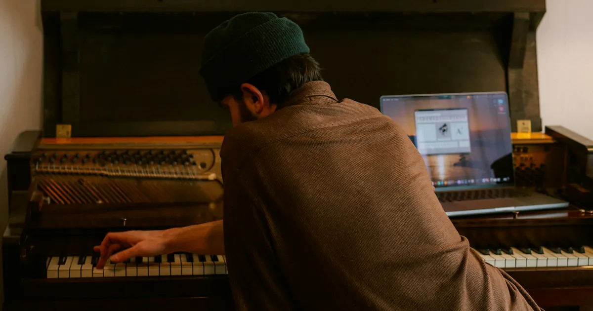 Músico trabalha em um piano com um notebook aberto ao lado.