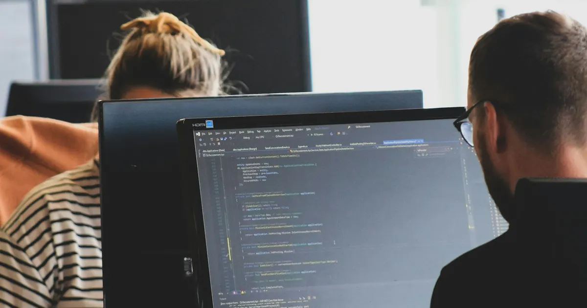 Duas pessoas trabalham com código em computadores no escritório.