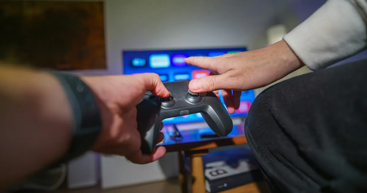 Mãos seguram um controle de videogame diante da televisão.