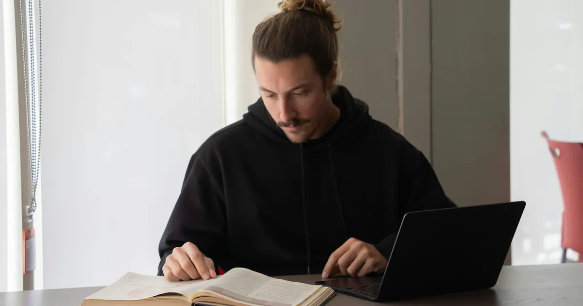 Pessoa consulta um livro ao mesmo tempo que utiliza um notebook.