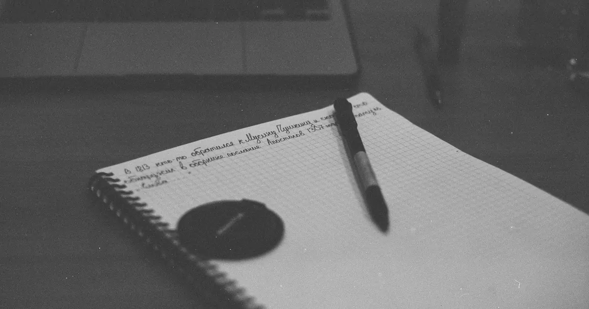 Caderno aberto e caneta sobre a mesa ao lado de um computador.